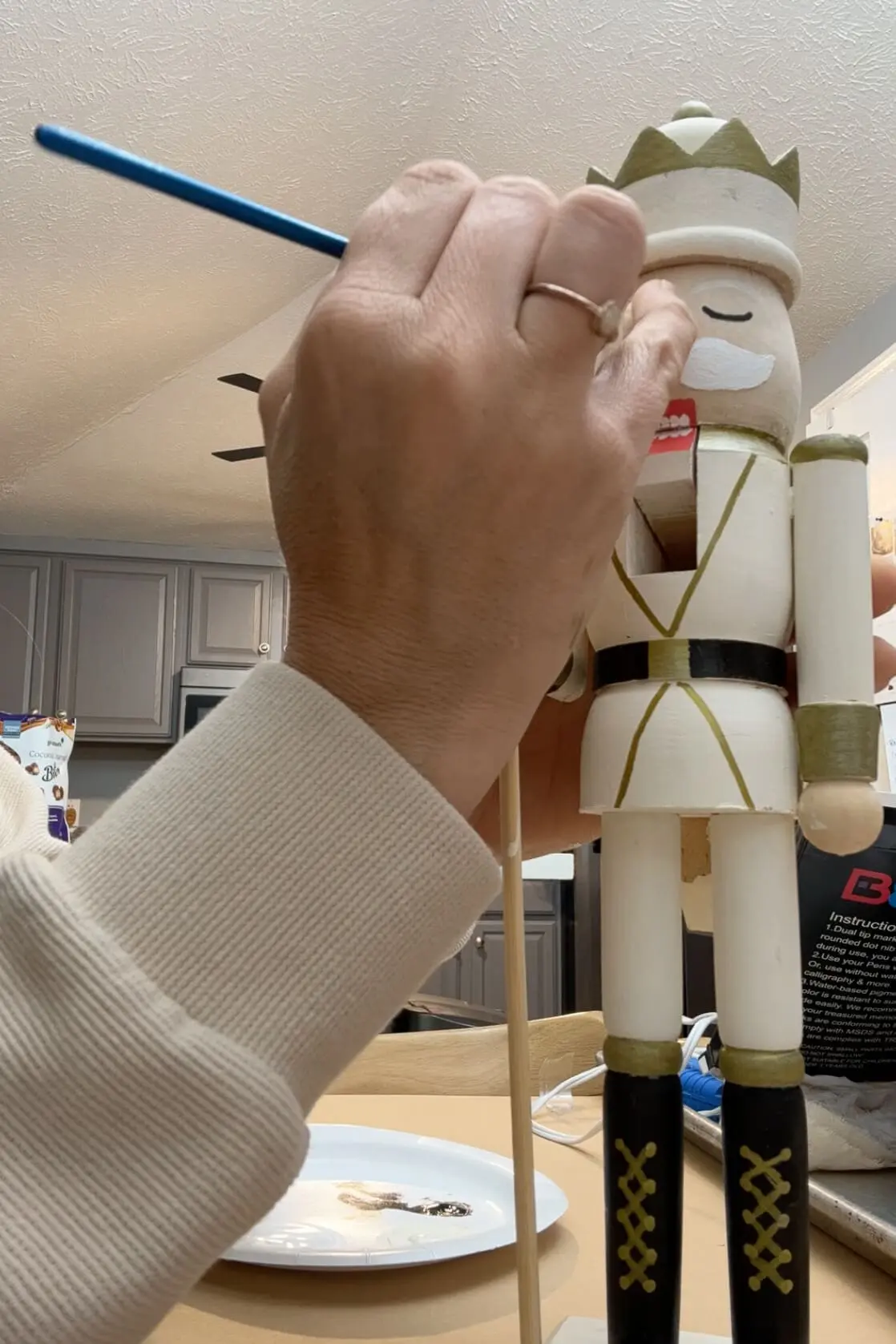Close-up of a hand painting details on the face of a wooden nutcracker, adding finishing touches to the mouth and mustache. Paints and brushes are visible in the foreground, adding a hands-on, artistic vibe to the scene.